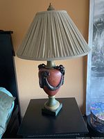 Photo of one porcelain lamp with gargoyle decoration and pleated linen shade on black side table next to bed.