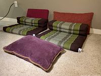 Photo of two meditation cushions with matching green and brown covers, two large red and rust rectangular pillows behind, and a smaller purple pillow on carpeted floor.