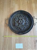 Full top-down view of the round metal decorative wall hanging showing the floral design, antiqued finish, and handles with measuring tape for scale.