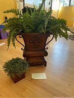 Photo of two silk plants: a large faux fern in an ornate brown urn planter and a smaller bush in a square brown pot on a wooden floor near a window.