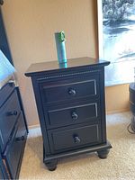 Black 3 drawer bedside table with round knobs, showing signs of scratches and wear. Dimensions approximately 19" wide, 18" deep, 27.5" tall.