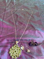 Photo showing two silver chain necklaces, one featuring a large green rhinestone pendant, alongside two pairs of earrings, one pair with silver heart-shaped charms and the other with purple bobble charms, all laid out on a dark textured fabric.