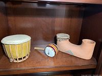 Shelved view of three small drums: yellow wooden drum, colorful rattle drum, ceramic boot-shaped drum