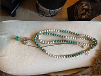 Turquoise and white stone mala necklace laid out on a white surface showing alternating beads, silver Ganesh locket and turquoise prayer wheel attached.