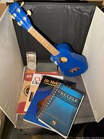 Blue soprano ukulele with tuning keys, metronome, and multiple instructional and song books arranged on a gray chair against a black background
