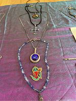 Photo showing five necklaces and three pendants laid on purple fabric, highlighting details of rhinestones, beads and enamel designs.