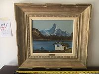 Framed vintage original oil painting on board featuring a mountain, lake, forest, and cabin, signed Alex, with measuring tape showing frame size