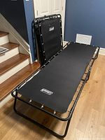First cot set up flat on floor with one cot folded and standing upright in background. Black fabric and metal frame with Coleman branding visible on fabric surface.
