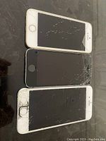 Three Apple iPhones placed front side up showing cracked and damaged screens, different colors including white/gold, black, and white/silver. Front screens heavily damaged.