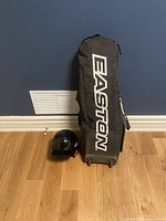 Whole lot showing Easton brand black rolling baseball bag upright next to a black batting helmet on floor.
