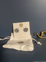 Pair of Rebekah Price flower-shaped clear stone stud earrings on white branded card with white drawstring pouch beneath