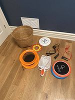 Assortment of outdoor game items including frisbee discs, orange mesh hoops, skipping ropes, white net paddle, and woven basket on wooden floor near a dark wall