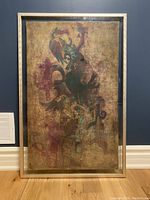 Full front view of the framed batik cloth artwork against a blue wall and wooden floor, showing details of the abstract figure and metallic frame.