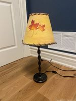 Full view of the lamp showing the yellow lampshade with printed leaf design and brown stitched edges, atop a dark twisted metal base on wooden floor next to blue wall and white door.