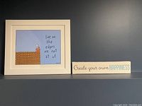 Framed print showing cartoon figure on cliff edge with inspirational text, next to a rectangular canvas with motivational slogan.