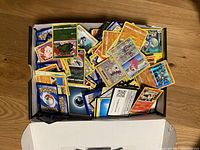 Shoebox filled with loose Pokémon and other character cards on a wooden floor