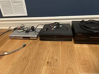 Three DVD/VCR players arranged side by side on wooden floor with wiring scattered.