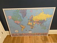 World map print mounted on hardboard leaning against a wall, showing full view of the map and the border with flags at bottom.