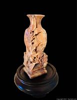 Front view of the carved stone vase mounted on a wooden stand, showing floral carved design on square base with flared rim.
