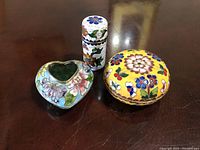 Photo showing all three cloisonne enamel items together on a dark surface: heart-shaped box, round yellow box, small cylindrical container with floral patterns.