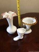 Three vintage Fenton glass pieces displayed on a wooden table with a measuring tape behind one tall vase. Items include two ruffled edged vases and one small hobnail boot figurine.