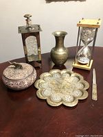 Photo showing six vintage decorative items: the brass and glass decanter with cracked glass panel; brass scalloped tray; brass vase; hourglass; lidded elephant trinket dish; envelope opener.