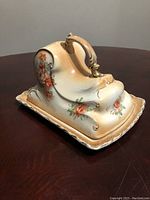 Side view of the covered cheese dish showing floral patterns and ornate handle on the cream porcelain base with visible crazing.