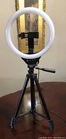 Ring light mounted on tripod stand against wall with framed picture in background