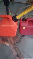 Photo showing two red gasoline fuel tanks on a glass outdoor table, one with a yellow flexible spout attached to the opening.