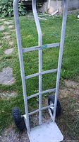 Front and side view of silver metal Haussmann dolly hand truck with two black pneumatic tires on grass background. One tire is flat, shown in close up.