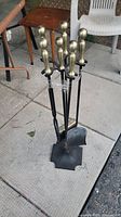 Full view of the six-piece fireplace tool set standing on floor, including shovel, broom, poker, and brass-handled tools with black shafts on metal stand.