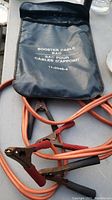 Black carrying bag labeled 'Booster Cable Sac Pour Cables D'appoint 11-2046-4' with orange booster cables and clamps.