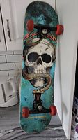 Front view of the skateboard showing skull and snake graphic on the deck with attached red wheels and black trucks