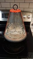 Front view of the vintage cone-shaped electric heater placed on a stove, showing metal mesh grill, orange top, and ring handle.