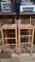 Two wicker and wood bar stools shown front view, highlighting square woven seats and wood legs with curved stylistic elements.