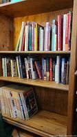 Three shelves packed with hardcover and softcover books. Shelves made of pine wood. Books include various genres and authors.