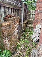 Stacks of various size bricks lined along a wooden fence and brick wall outdoors with some weeds around.