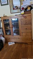Front view of wood cabinet showing two glass panel doors and five drawers with wood knobs, carved front detail, and surrounding room environment.
