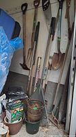 Various garden tools arranged against a wall including shovels, rakes, pitch fork, spades, and plant containers on the floor, some rust and dirt visible indicating use.