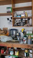 Photo of shelves and workbench containing various paint cans, spray wax, caulk, tools, screws, and hardware items in containers and boxes.