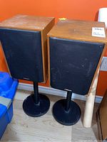 Pair of Klipsch speakers on black metal stands with circular bases, positioned against an orange wall, showing wooden cabinet tops and black fabric grills.