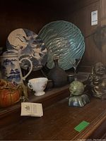 Shelf display showing large green textured plate, blue and white crane plate, pumpkin teapot, Buddha statue, and green sphere on base.