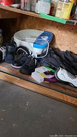 Photo showing various sports and fitness items including foot spa, black Asham shoes, shin guards, and boxed yoga knee pads, all arranged on floor against shelving.