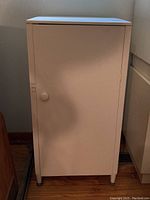 Closed view of white painted wood storage cabinet with round knob and four legs.