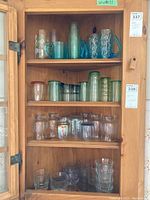 Full view of four shelves containing glasses of different colors including green, blue, and clear. Includes mugs, tumblers, and other glass types.