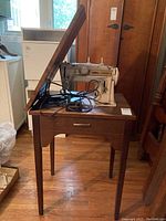 Singer sewing machine mounted inside wooden cabinet table with top lid open showing sewing machine and power cord.