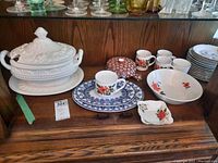 Shelf display showing white ceramic soup tureen with lid, blue and white oval plates, Sadler mugs, beige bowl, and Royal Albert side plate.