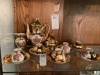 Full set of gold-toned tea set on wooden shelf, includes teapot, cream pitcher, sugar bowl with lid, six tea cups, and six saucers, decorated with pink rose and couple scenes.