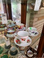 Display of five floral bone china teacups with matching saucers in glass cabinet, showing variety of patterns and colors, some with gold trim