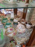 Glass cabinet shelf displaying an assortment of six teacups and saucers in various floral and gold designs with minor wear observed